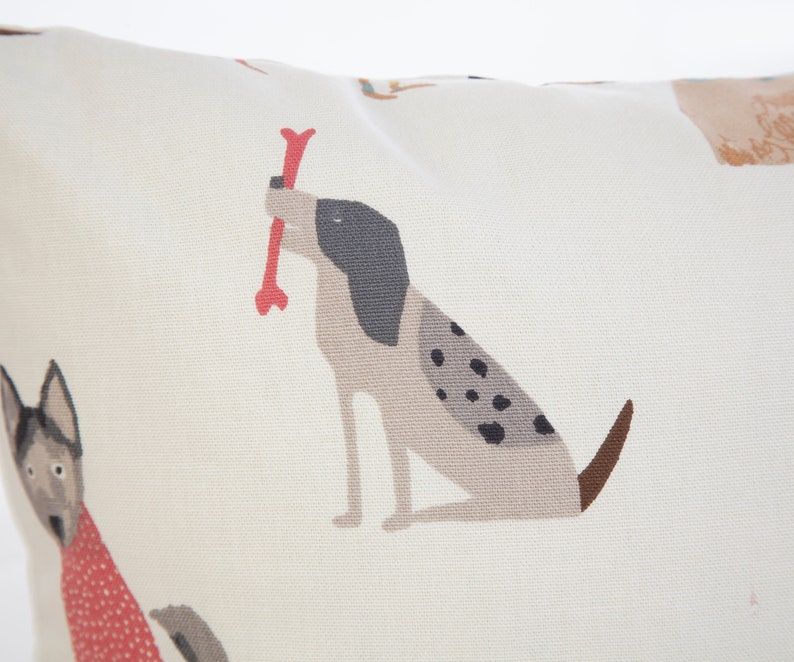 K&ouml;nnte beinhalten: Ein wei&szlig;er Kissenbezug mit einem sich wiederholenden Muster aus Hunden. Die Hunde sind im Cartoon-Stil illustriert und sind verschiedener Rassen. Die Hunde sind in Braun-, Schwarz- und Wei&szlig;t&ouml;nen gehalten. Ein Hund h&auml;lt einen roten Knochen im Maul.