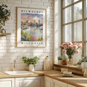 A framed Milwaukee, Wisconsin watercolor cityscape print hanging on a white brick wall in a bright kitchen, surrounded by greenery and soft decor.