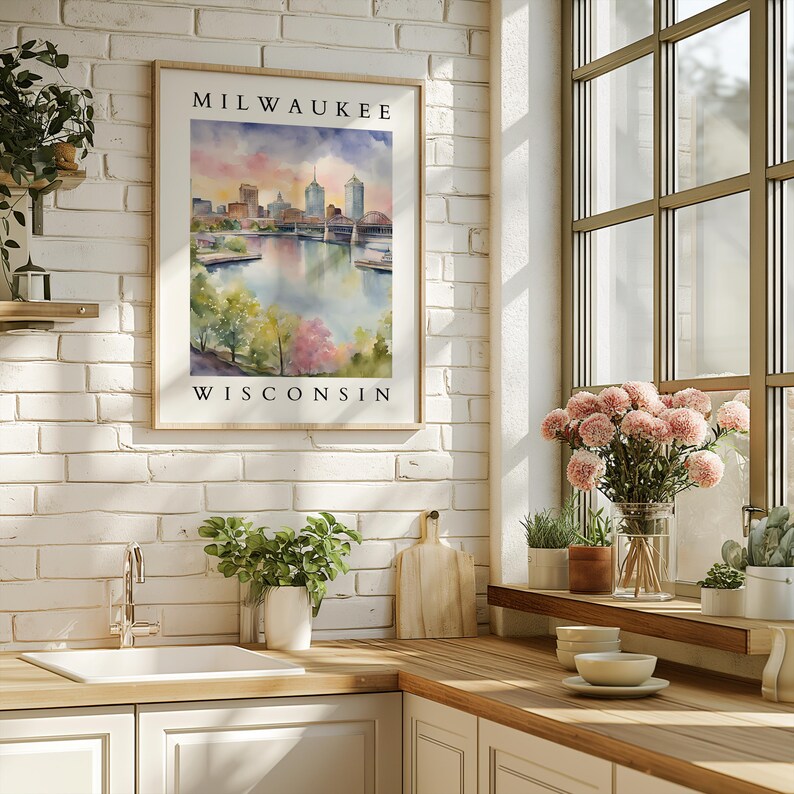 A framed Milwaukee, Wisconsin watercolor cityscape print hanging on a white brick wall in a bright kitchen, surrounded by greenery and soft decor.