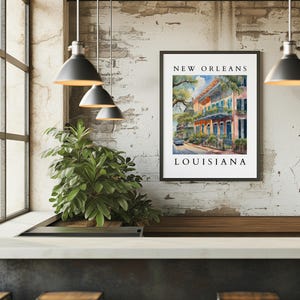 May include: A framed watercolor painting of a street scene in New Orleans, Louisiana. The painting depicts colorful buildings with balconies and lush greenery. The text "NEW ORLEANS" and "LOUISIANA" is printed below the image.