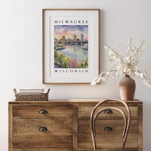 A Milwaukee, Wisconsin cityscape print in a wooden frame, elegantly placed above a rustic wooden dresser with white floral decor and a woven chair.