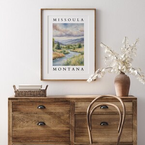 May include: A framed watercolor painting of a river winding through a valley with mountains in the background. The painting is titled "Missoula, Montana".