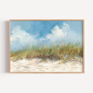 May include: Watercolor painting of a beach scene with a blue sky and fluffy white clouds. Green sea grass grows on a sandy dune. The artwork is framed in a light-colored wooden frame, creating a serene coastal landscape.