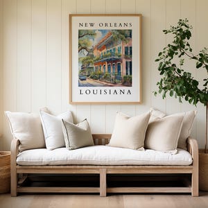 May include: A white linen bench with pillows sits in front of a white wall with a framed print that reads "NEW ORLEANS LOUISIANA". A potted plant with green leaves is on the right side of the image.