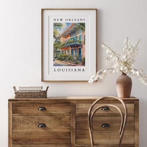 May include: A framed watercolor painting of a colorful building in New Orleans, Louisiana. The building has a balcony and is painted in shades of orange, yellow, and blue. The painting is framed in a light wood frame.