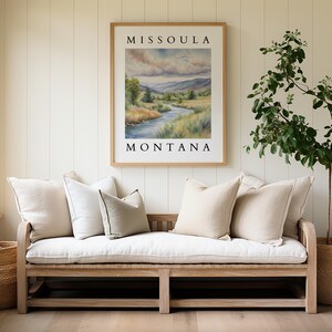 May include: A white wooden bench with a white cushion and several throw pillows. The bench is in front of a white wall with a framed print that reads "Missoula Montana" and a potted plant.