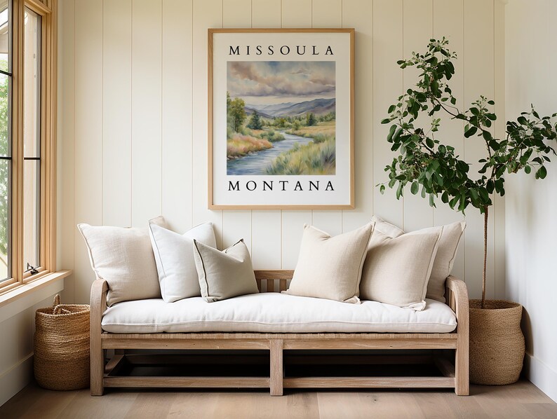 May include: A white wooden bench with a white cushion and several throw pillows. The bench is in front of a white wall with a framed print that reads "Missoula Montana" and a potted plant.