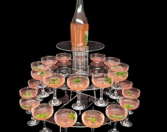 Puede incluir: Un soporte de acrílico de varios niveles con una botella de champán rosado y 15 copas de champán transparentes llenas de una bebida rosada. Las copas están dispuestas en forma circular en el soporte.