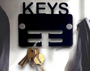 May include: Black metal key holder with the word "KEYS" in large letters. The holder has three slots for keys and two silver dots on the sides. A set of keys is hanging from the holder.