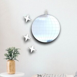 May include: A silver disco ball mirror with a white background. There are three white star-shaped mirrors on the wall around the disco ball.