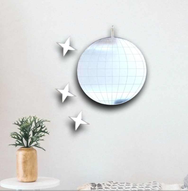 May include: A silver disco ball mirror with a white background. There are three white star-shaped mirrors on the wall around the disco ball.
