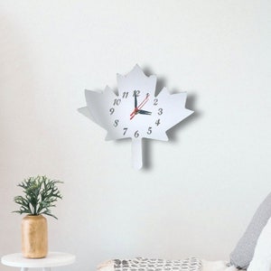 Peut inclure: Une horloge murale en forme de feuille d'érable blanche avec des chiffres et des aiguilles noirs. L'horloge est montée sur un mur blanc.