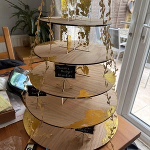 May include: A four-tiered wooden cake stand with gold leaf detailing and a decorative vine. Each tier has a small chalkboard sign. The stand is designed to display treats like cookies and blondies. The stand is made of wood.