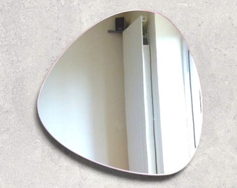 Peut inclure: Un grand miroir de forme ovale et libre, avec une fine bordure rose. Le miroir reflète un mur blanc et une porte blanche.
