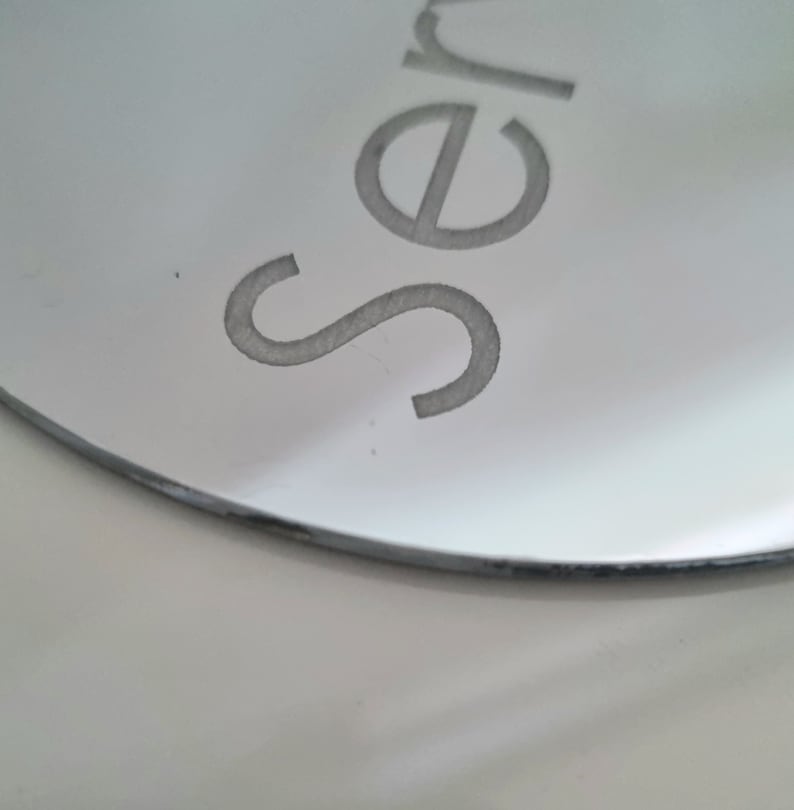 May include: A close-up of a round, silver mirror with the word "Ser" etched into the surface.
