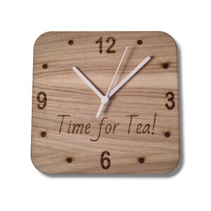 Könnte beinhalten: Eine quadratische Holzuhr mit weißen Zeigern und schwarzen Zahlen. Das Zifferblatt der Uhr trägt den Text "Time for Tea!" eingraviert.