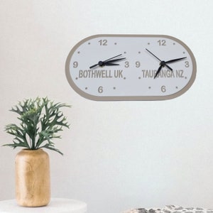 Peut inclure: Une horloge ovale blanche avec des aiguilles noires et deux fuseaux horaires affichés. L'horloge indique "Bothwell UK" et "Tauranga NZ".