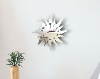 May include: A silver mirrored clock shaped like a sun with black numbers and hands. The clock is mounted on a white wall.