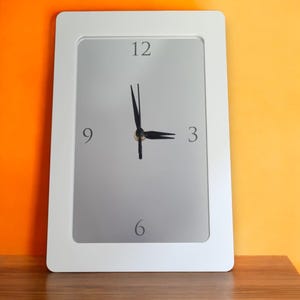 May include: White framed clock with black hands and grey face. The clock face has numbers 12, 9, 3, and 6.