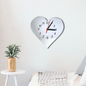 Op de afbeelding: Een witte, hartvormige wandklok met zwarte cijfers en wijzers. De secondewijzer is rood. De klok is aan een witte muur bevestigd. Een kleine plant in een houten vaas staat op een witte bijzettafel.