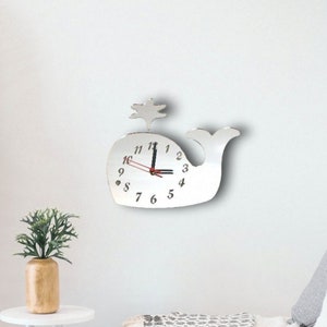 Könnte beinhalten: Eine weiße, walförmige Uhr mit schwarzen Zeigern und roten Stundenmarkierungen. Die Uhr hat einen kleinen weißen Baum oben drauf und ist an einer weißen Wand montiert.