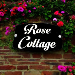 Könnte beinhalten: Ein schwarzes Schild mit weißen Buchstaben, auf dem "Rose Cottage" steht, ist an einer Backsteinmauer befestigt. Das Schild ist von rosa Blumen und grünem Laub umgeben.