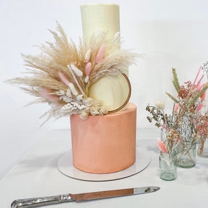 Könnte beinhalten: Eine zweistöckige Hochzeitstorte mit einer weißen unteren und einer pfirsichfarbenen oberen Etage. Die Torte ist mit einem Holzring und einem Strauß getrockneter Pampasgräser und rosa Blumen verziert.