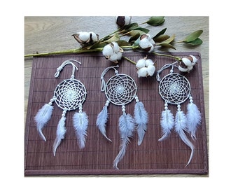 May include: Three white dreamcatchers with white feathers hanging from a brown bamboo mat. The dreamcatchers have a white beaded border and a white woven web.
