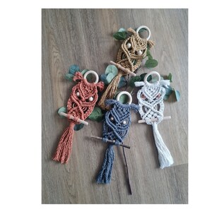 Könnte beinhalten: Fünf Makramee-Eulen-Wandbehänge mit Holzringen und Perlen. Die Eulen sind in verschiedenen Farben, darunter Braun, Orange, Grau und Weiß.