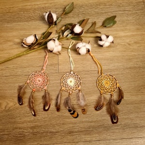 May include: Three dreamcatchers with brown feathers and a variety of colored beads. The dreamcatchers are hanging from a string and are against a wooden background.