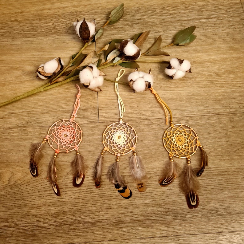 May include: Three dreamcatchers with brown feathers and a variety of colored beads. The dreamcatchers are hanging from a string and are against a wooden background.