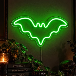 May include: A bright green neon sign shaped like a bat with outstretched wings. The sign emits a vibrant glow against a dark wall. Below the sign are stacked books labeled Witchcraft, Dark Aesthetic, and Halloween, a small pumpkin, and a skull.
