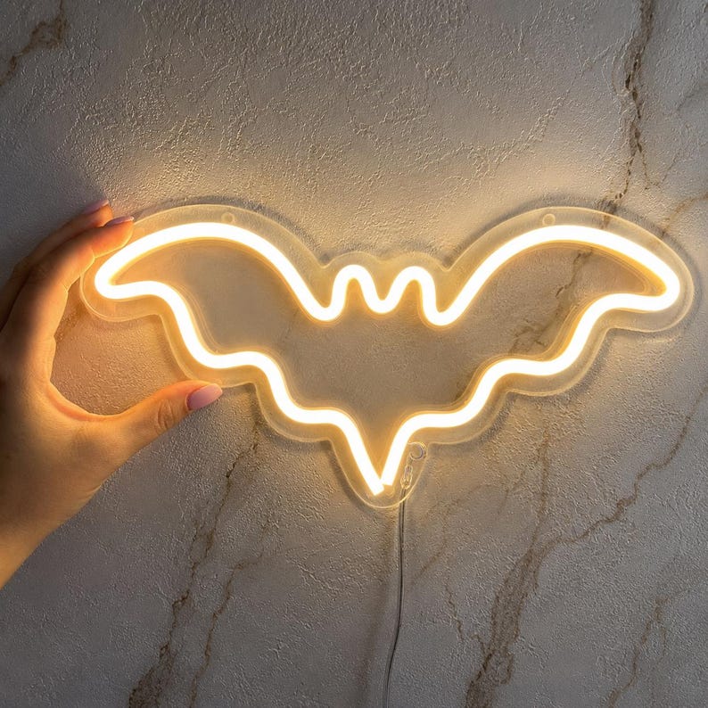 May include: A bat-shaped neon sign emitting a warm yellow glow. The light is mounted on a clear acrylic backing that follows the outline of the bat silhouette. A thin power cord extends from the bottom of the fixture, which is illuminated.