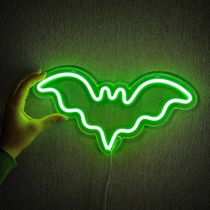 May include: A bright neon sign shaped like a bat, emitting a vibrant green light. The bat silhouette features curved wings and a pointed bottom edge, mounted on a clear acrylic backing with a thin power cord extending from the bottom center.