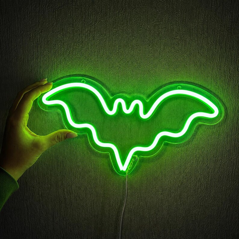 May include: A bright neon sign shaped like a bat, emitting a vibrant green light. The bat silhouette features curved wings and a pointed bottom edge, mounted on a clear acrylic backing with a thin power cord extending from the bottom center.