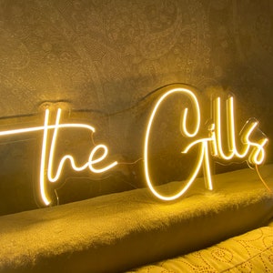 K&ouml;nnte beinhalten: Ein Neon-Schild in warmem Gelb zeigt die Worte "the Gills" in einer Schreibschrift. Das Schild ist an einer Wand befestigt und eingesteckt, wobei die beleuchteten Buchstaben ein sanftes Leuchten erzeugen.
