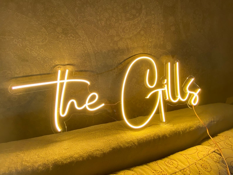 K&ouml;nnte beinhalten: Ein Neon-Schild in warmem Gelb zeigt die Worte "the Gills" in einer Schreibschrift. Das Schild ist an einer Wand befestigt und eingesteckt, wobei die beleuchteten Buchstaben ein sanftes Leuchten erzeugen.