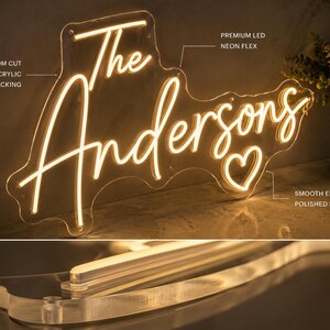 K&ouml;nnte beinhalten: Neon-Schild mit dem Schriftzug "The Andersons" in geschwungener Schrift, hergestellt aus hochwertigem LED-Neon-Flex und auf klarem Acryl montiert. Das Design beinhaltet ein Herzsymbol und hat glatte, polierte Kanten.