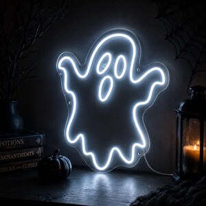 May include: A ghost-shaped neon sign emitting a bright white light. The sign features a classic cartoon ghost silhouette with two oval eyes and an oval mouth, mounted on a clear acrylic backing with visible mounting holes along the edges.