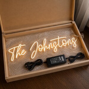 K&ouml;nnte beinhalten: Ein Neonschild in einer Holzkiste mit der Aufschrift "The Johnstons" in einer kursiven Schriftart. Das Schild leuchtet mit einem warmen, sanften Schein. Ein schwarzes Netzteil und Kabel sind sichtbar.
