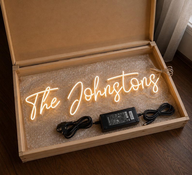 K&ouml;nnte beinhalten: Ein Neonschild in einer Holzkiste mit der Aufschrift "The Johnstons" in einer kursiven Schriftart. Das Schild leuchtet mit einem warmen, sanften Schein. Ein schwarzes Netzteil und Kabel sind sichtbar.