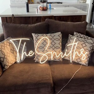K&ouml;nnte beinhalten: Ein braunes Sofa mit dekorativen Kissen und einem Neonschild mit der Aufschrift "The Smiths" in Schreibschrift. Die Kissen haben ein geometrisches Muster in Grau- und Wei&szlig;t&ouml;nen. Das Neonschild leuchtet in warmwei&szlig;em Licht.