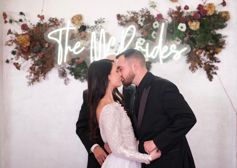 K&ouml;nnte beinhalten: Ein Brautpaar k&uuml;sst sich bei einer Hochzeitszeremonie. Die Braut tr&auml;gt ein wei&szlig;es Kleid mit langen &Auml;rmeln, der Br&auml;utigam einen schwarzen Smoking. Ein Neonschild mit der Aufschrift "The McBrides" befindet sich &uuml;ber ihnen, umgeben von Blumenschmuck.