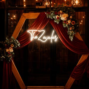 May include: A wooden hexagonal wedding arch decorated with burgundy velvet fabric and floral arrangements. The arch features a neon sign that reads "The Zayafas". The background is dark, with warm lighting.