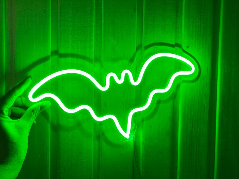 May include: A bright green neon sign shaped like a bat with outstretched wings. The light emits a vibrant glow, highlighting the distinct silhouette of the creature against the vertical paneling. The sign features a clear acrylic backing.