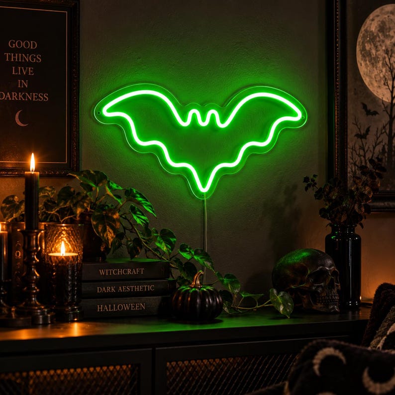 May include: A bright green neon sign shaped like a bat with outstretched wings. The sign emits a vibrant glow against a dark wall. Below the sign are stacked books labeled Witchcraft, Dark Aesthetic, and Halloween, a small pumpkin, and a skull.