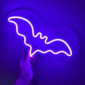 May include: A bright neon sign shaped like a bat, emitting a vibrant purple and white glow. The bat silhouette features curved wings and a distinct head shape, illuminated by a continuous light tube. A thin power cord hangs from the bottom edge.