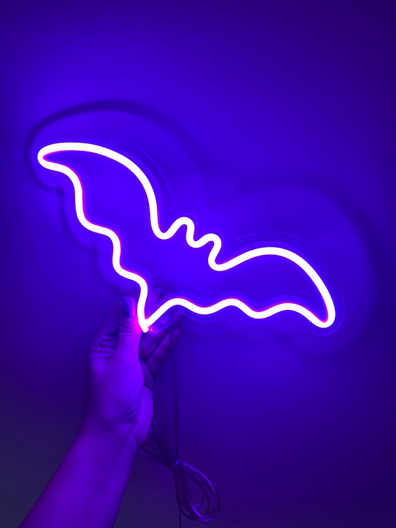 May include: A bright neon sign shaped like a bat, emitting a vibrant purple and white glow. The bat silhouette features curved wings and a distinct head shape, illuminated by a continuous light tube. A thin power cord hangs from the bottom edge.