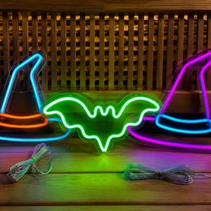 May include: Three neon signs featuring two witch hats and a bat. The left hat is blue with an orange band, the center bat is bright green, and the right hat is purple with a blue band. Each sign has a clear power cord attached to the base.