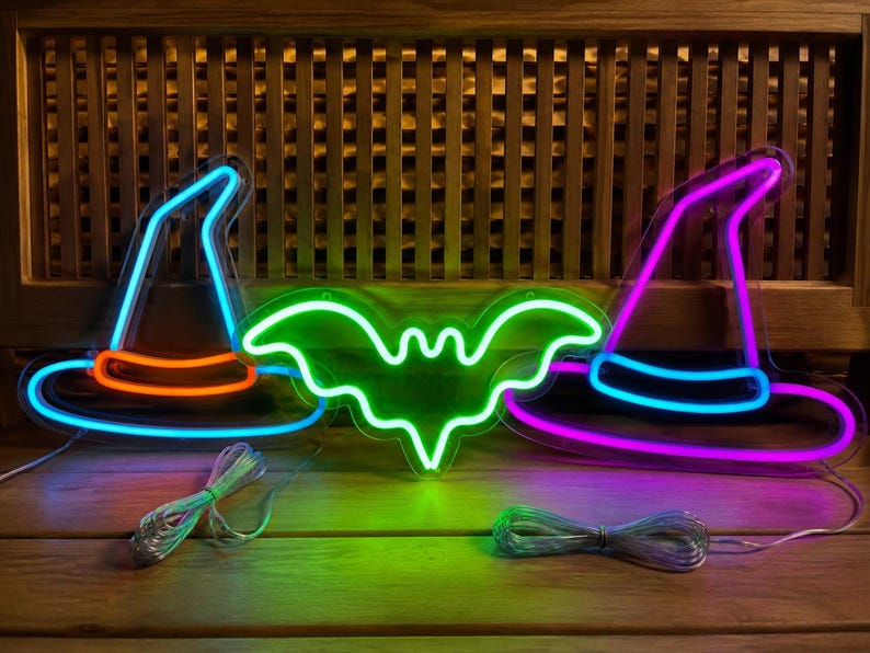 May include: Three neon signs featuring two witch hats and a bat. The left hat is blue with an orange band, the center bat is bright green, and the right hat is purple with a blue band. Each sign has a clear power cord attached to the base.